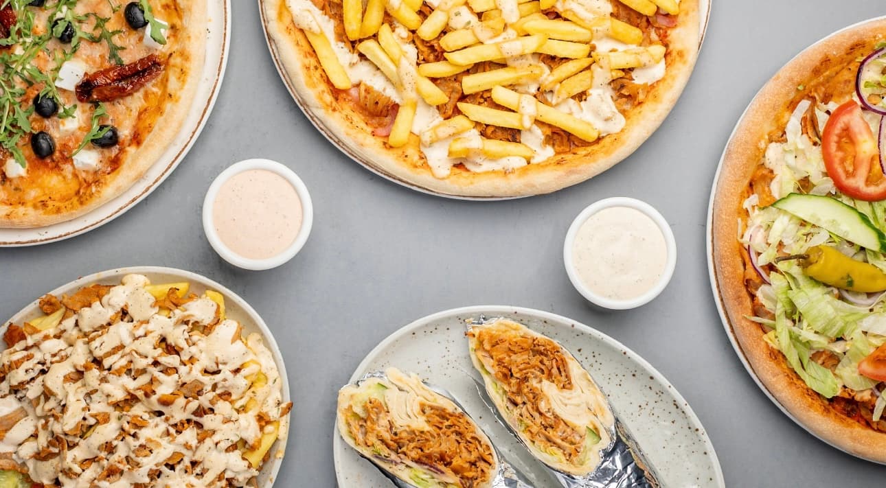 Assorted kebab pizzas, wraps, and loaded fries with dipping sauces on a grey surface.
