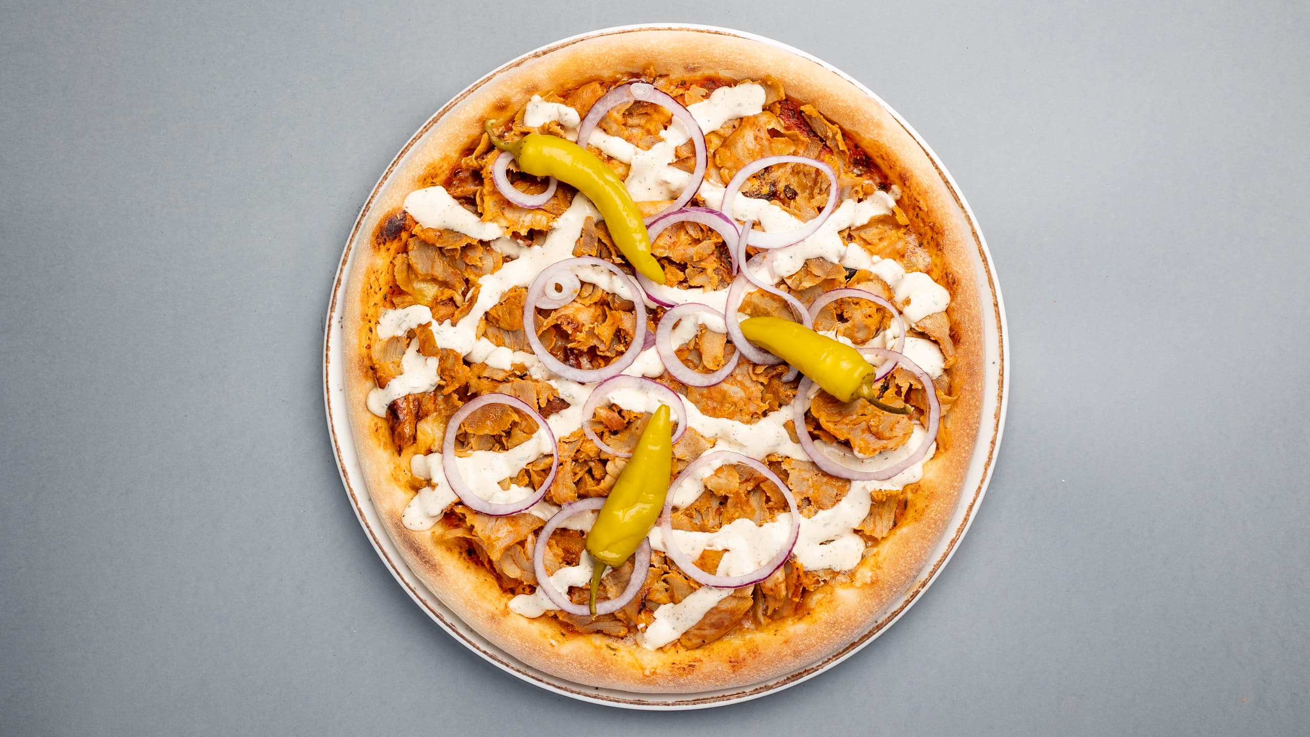 Kebab pizza topped with red onions, white sauce, and green chilies on grey background.