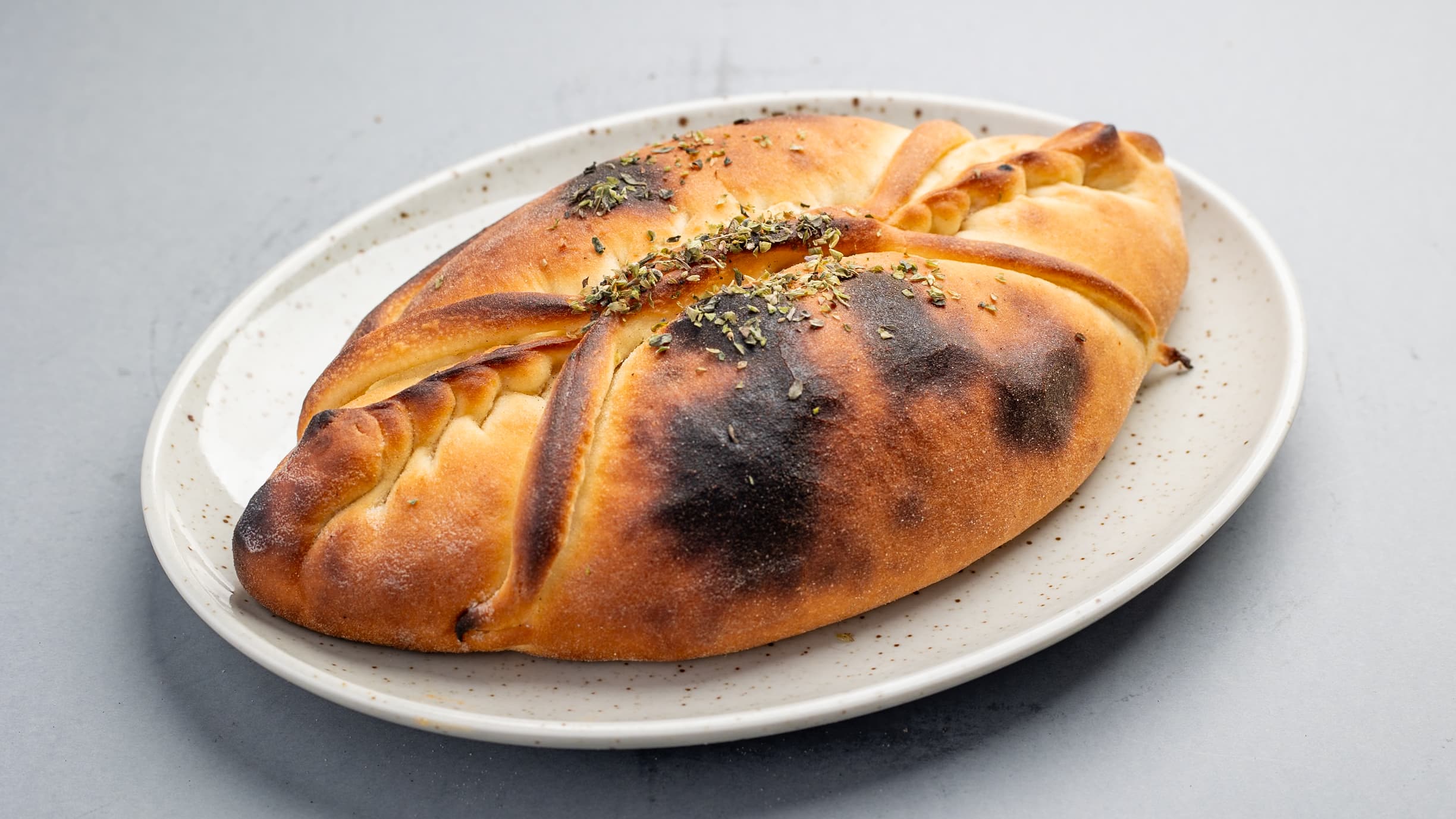 Golden-brown calzone with charred crust and dried herbs served on a speckled white plate.