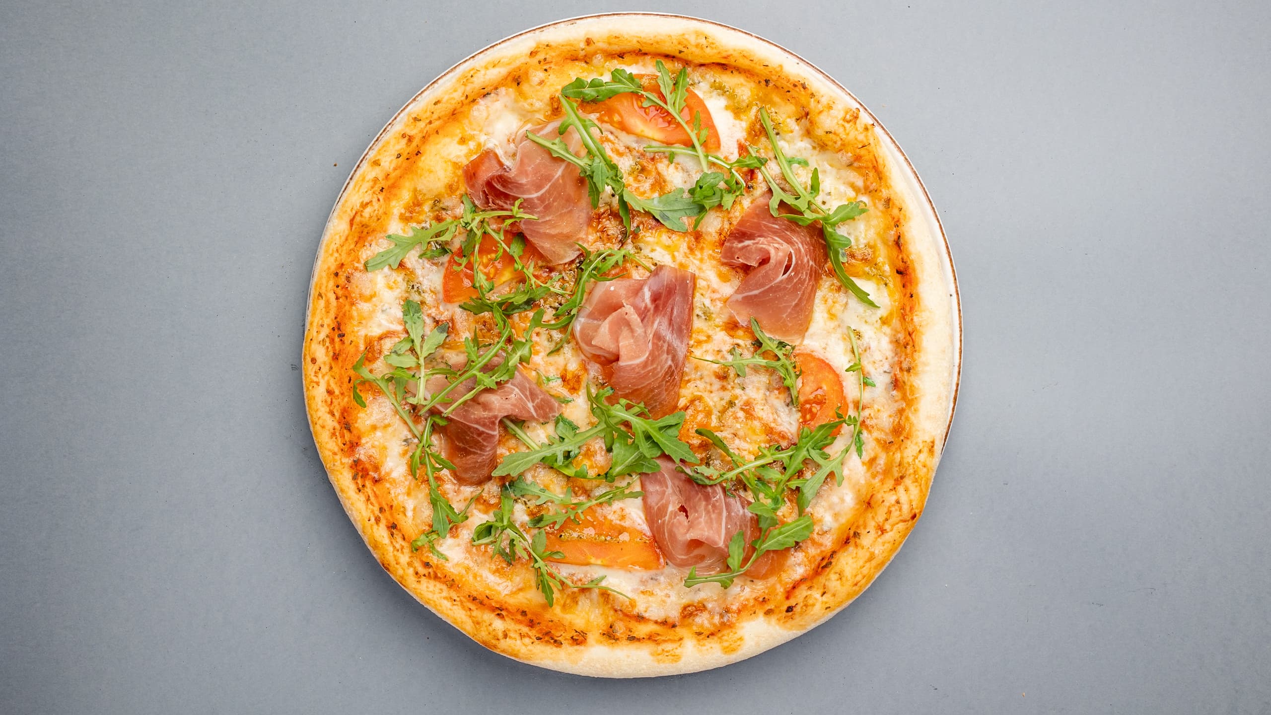 Thin-crust pizza topped with prosciutto, fresh arugula, and tomato slices on a gray background.