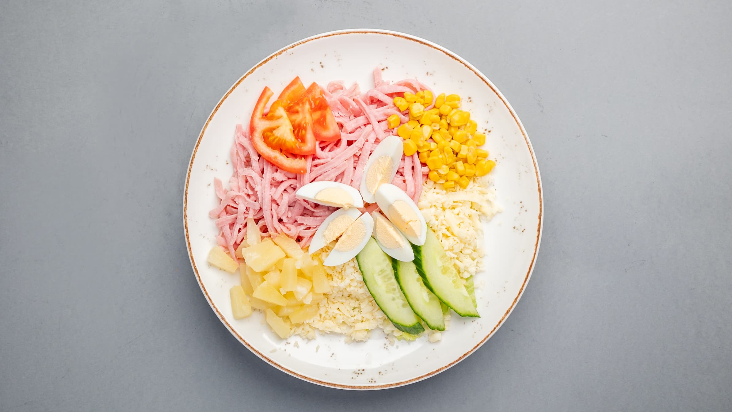 Plate of shredded ham, corn, cheese, pineapple, cucumber, tomato, and hard-boiled egg wedges.