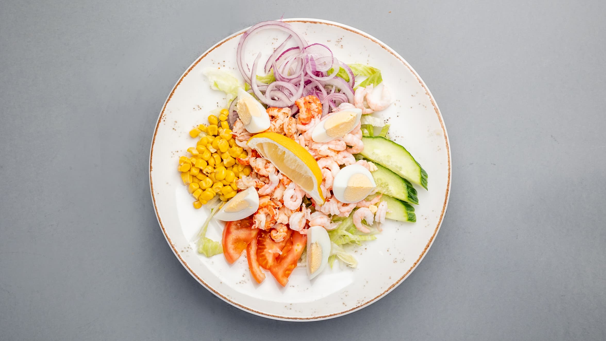 Shrimp salad with eggs, corn, red onions, cucumber, tomato, and lemon on a white plate.
